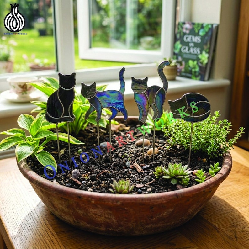 May include: Four stained glass cat garden stakes are inserted into a large, round, terracotta-colored planter filled with soil and various green plants. The cats feature iridescent, blue, purple, and black glass panels with thin metal solder lines.