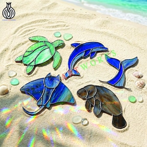 May include: A collection of five stained glass sea creatures including a green sea turtle, a blue dolphin, a blue whale tail, a blue stingray, and a brown manatee, all featuring dark metal soldering lines and light-refracting glass textures.