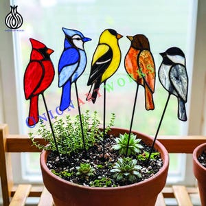 Könnte beinhalten: Fünf Buntglasvögel auf dünnen Metallstäben, darunter ein roter Kardinal, ein Blauhäher, ein gelber Stieglitz, ein orangefarbener Vogel und eine schwarz-weiße Meise, angeordnet in einem Terrakottatopf mit kleinen grünen Sukkulenten.