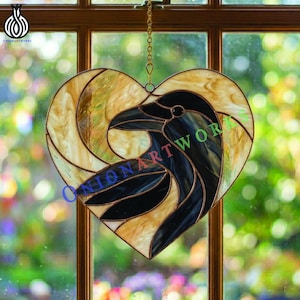May include: Stained glass heart-shaped artwork featuring a raven design. The raven is depicted in black and gray, set against a background of amber and gold glass. The artwork is suspended by a gold chain and is displayed in front of a window.