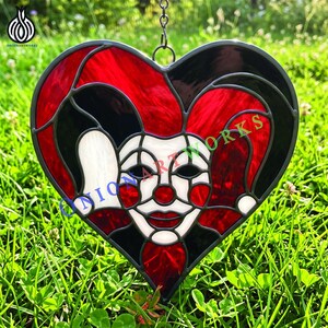 Puede incluir: Un atrapasoles de vidrieras en forma de corazón con un diseño de cara de bufón en piezas de vidrio rojo, negro y blanco unidas por líneas de soldadura de metal oscuro. El bufón tiene nariz roja, cara blanca y gorro rojo y negro. El texto dice ONIONARTWORKS.