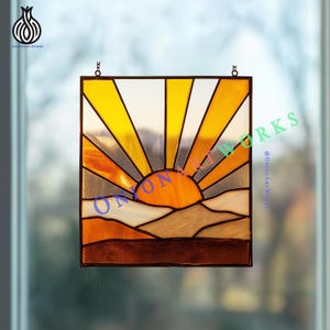 May include: Stained glass panel depicting a sunset scene with a vibrant orange sun and radiating yellow rays. The design includes layered mountain shapes in shades of brown and tan. The panel is framed in black and hangs from a chain.