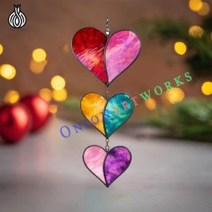 May include: A hanging stained glass heart decoration. The decoration features three hearts in shades of red, pink, yellow, teal, and purple. Each heart is divided into two sections with different colours. The hearts are connected by metal chains.