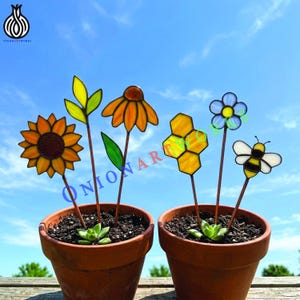 Puede incluir: Estacas de jardín de vidrieras de colores con forma de girasol, hojas, una margarita, un panal, una flor azul y una abeja. Las estacas están en macetas de terracota con suculentas, sobre un cielo azul.
