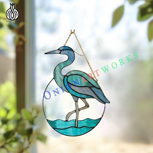 May include: Stained glass heron suncatcher in shades of blue and teal, with a gold chain for hanging. The heron is depicted standing in water within an oval frame. The text "ONION ARTWORKS" is visible on the glass.