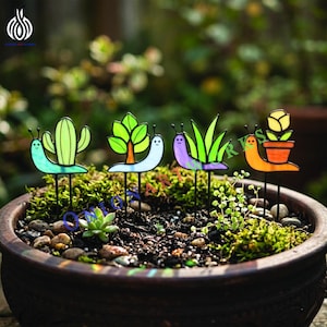 Puede incluir: Un conjunto de estacas de jardín coloridas con diseños caprichosos de caracoles. Cada estaca muestra un elemento diferente: un cactus, un árbol, hierba y una flor en una maceta. Las estacas están colocadas en una maceta llena de tierra y vegetación.