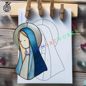 Può includere: Un'opera d'arte in vetro colorato raffigurante una figura religiosa con velo blu e aureola, appesa davanti a un modello numerato. L'opera è fissata a una tavola di legno con mollette. Il logo "ONION ARTWORKS" è visibile.
