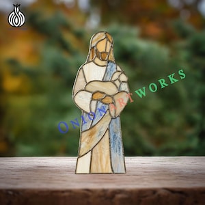 Könnte beinhalten: Glasmalerei-Figur von Jesus, der ein Lamm hält. Die Figur hat einen hellbraunen Bart und Haare und trägt ein weißes Gewand mit blauen und beigen Akzenten. Die Figur steht auf einer Holzoberfläche.