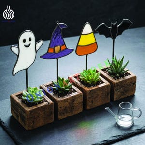 May include: Four square wooden planters containing small green succulents, each featuring a stained glass topper: a white ghost, a purple witch hat, a yellow and orange candy corn, and a black bat. A small clear glass watering can sits nearby.