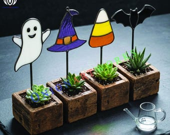 May include: Four square wooden planters containing small green succulents, each featuring a stained glass topper: a white ghost, a purple witch hat, a yellow and orange candy corn, and a black bat. A small clear glass watering can sits nearby.