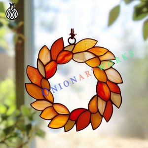 Op de afbeelding: Een ronde glas-in-lood krans met individuele bladvormige stukjes in tinten oranje, rood, geel en amber. De glazen segmenten zijn verbonden door donker metaalsoldeer en een kleine metalen ring aan de bovenkant maakt het ophangen mogelijk.