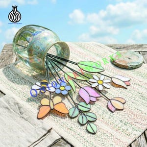 Puede incluir: Un jarrón de vidrio con estacas de jardín decorativas de vidrieras. Las estacas representan flores, hojas y una mariposa en tonos rosa, azul, amarillo y verde. El jarrón tiene un acabado iridiscente, y la escena es al aire libre.