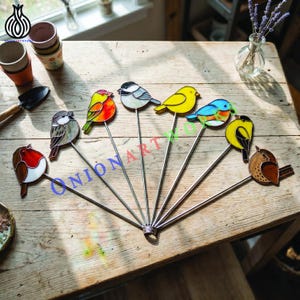 Pode incluir: Uma coleção de oito ornamentos de pássaros em vitral sobre hastes de metal, apresentando várias cores, incluindo vermelho, amarelo, azul e marrom. Cada pássaro é feito com segmentos de vidro distintos e linhas de chumbo em leque.