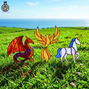 Op de afbeelding: Drie glas-in-lood figuren bestaande uit een rode en oranje draak, een gele en oranje feniks met gespreide vleugels, en een witte eenhoorn met blauwe manen en staart. Elk stuk is gemaakt met donkere metalen omlijningen en gekleurd glas.
