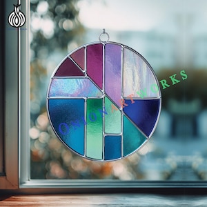 May include: A round stained glass window with a geometric design. The window features a variety of colors, including purple, blue, green, and red, and is framed with a silver metal border. The window is hanging in front of a window.