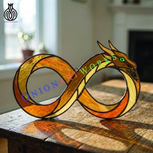 Könnte beinhalten: Glasfenster-Unendlichkeitszeichen mit einem Drachenkopf. Der Drache ist braun und goldfarben mit grünen Augen. Das Unendlichkeitszeichen besteht aus orangefarbenem, gelbem und goldenem Glas. Das Stück wird auf einer Holzoberfläche ausgestellt.
