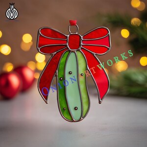 Puede incluir: Adorno navideño de vidriera con forma de lazo y pepinillo. El lazo es rojo, y el pepinillo es verde y verde claro con puntos negros. El adorno tiene un borde de metal plateado y una cinta roja para colgar. El texto "ONION ARTWORKS" es visible.