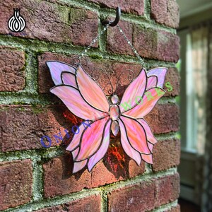 Könnte beinhalten: Ein Buntglas-Schmetterling in Rosa- und Orangetönen, der an einer Kette hängt. Das Kunstwerk befindet sich vor einer roten Backsteinmauer. Die Flügel haben ein geschichtetes Design, und der Körper hat einen zentralen Edelstein.