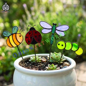 May include: A white ceramic pot filled with succulents, featuring four colorful stained-glass garden stakes: a bee, ladybug, dragonfly, and caterpillar. The stakes are in yellow, red, green, and purple. The text "ONIONARTWORKS" is visible.