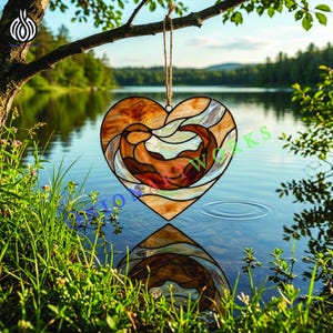 Pode incluir: Um apanhador de sol em vitral em forma de coração com um desenho de lontra, suspenso por um fio de barbante. O vidro apresenta tons de castanho, bege e creme, com um contorno preto. O apanhador de sol está pendurado em frente a um lago.