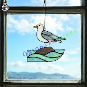 Puede incluir: Vidriera de gaviota con cuerpo blanco, alas grises y pico amarillo, posada sobre una roca marrón sobre olas azules y verdes. El texto "ONION ARTWORKS" es visible. Decoración para colgar delante de una ventana.