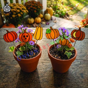 May include: Two terracotta pots containing small green plants and purple flowers, each decorated with multiple stained glass pumpkin stakes in orange and yellow hues, featuring various pumpkin shapes and a jack-o'-lantern face.