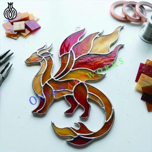 May include: A stained glass dragon suncatcher featuring a vibrant color palette of red, orange, and yellow glass pieces held together by silver solder lines. The dragon is depicted in a dynamic pose with wings spread and a long, curved tail.