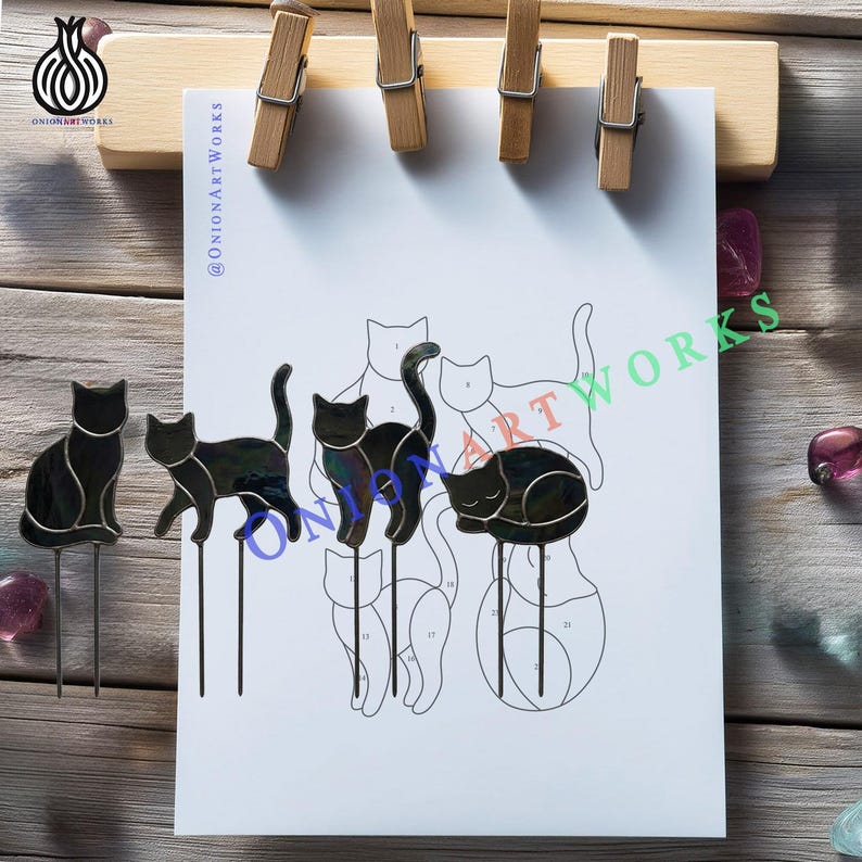 May include: Four black stained glass cat figures with metal stakes, displayed alongside a white paper pattern sheet featuring outlines of cats. The glass cats are in various poses, including sitting, walking, and sleeping, with visible solder lines.