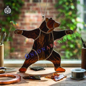 Pode incluir: Um urso de vitral com os braços estendidos, pendurado por uma corrente prateada. O urso apresenta tons de castanho e âmbar, com um aspeto texturizado. O texto "ONONAR WORKS" é visível. Um artigo decorativo.