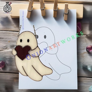 May include: A stained glass ghost holding a red heart, with black eyes and a chain for hanging. The ghost is cream-colored with a copper-colored outline. A line drawing of a ghost is behind it on a white paper, held by wooden clips.
