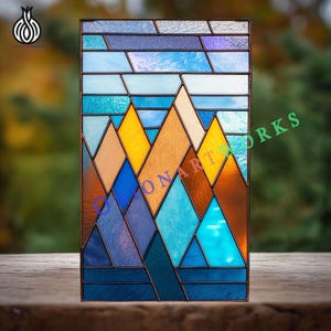 Puede incluir: Panel rectangular de vidriera con diseño de paisaje montañoso. La obra utiliza tonos de azul, amarillo y marrón, con el texto "ONARTWORKS" visible. Enmarcado con un borde metálico oscuro, descansa sobre una superficie de madera.