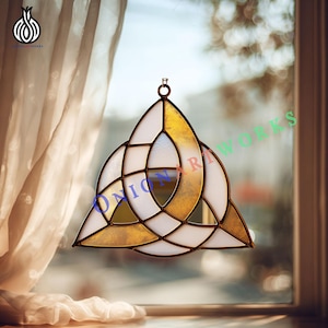 May include: A stained glass Celtic knot, featuring a gold and white color scheme. The design is a three-part knot, with a circular top for hanging. The text "ONION ARTWORKS" is visible on the glass. The piece is displayed in front of a window.