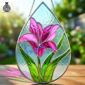 May include: Stained glass window art in a teardrop shape, featuring a pink lily with green leaves. The glass has a textured appearance and is framed with a silver metal border. The piece is suspended by a chain.