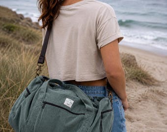 May include: A sage green corduroy duffel bag featuring a rectangular white fabric label with black text, dark brown trim, black zippers, and a detachable dark brown shoulder strap with antique brass-toned metal hardware clips.