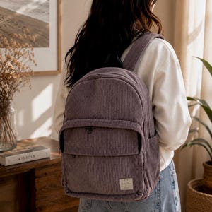 May include: A dusty purple corduroy backpack featuring a large main compartment, a smaller front zippered pocket, and a rectangular fabric patch with text on the lower right corner. The bag has matching shoulder straps and side pockets.