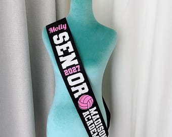 May include: A black sash featuring the name Molly in pink script, the word SENIOR in large white block letters, the year 2027 in pink, a pink volleyball graphic, and the text MADISON ACADEMY in white block letters, displayed on a teal mannequin.