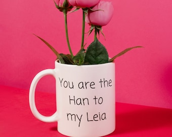 Może przedstawiać: Biały ceramiczny kubek z czarnym napisem "You are the Han to my Leia", z różowymi różami i pąkami. Kubek ma zakrzywiony uchwyt i stoi na czerwonej powierzchni na różowym tle.
