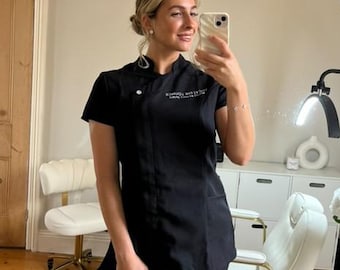 May include: A black uniform with short sleeves and a mandarin collar. The uniform has a side pocket and a logo on the chest. The person is holding a phone and taking a selfie in front of a mirror. The background includes a white chair and a white cabinet.
