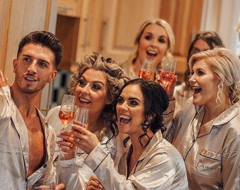 May include: A group of people wearing white satin pajamas are smiling and holding glasses of champagne. One person is taking a selfie with a smartphone.