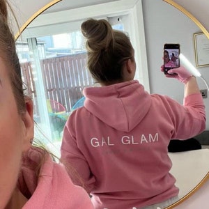 May include: A pink hoodie with the text "GAL GLAM" printed on the back. The hoodie is being worn by a person with their hair in a bun. The person is taking a selfie in a mirror.
