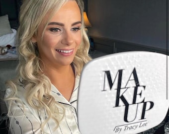 May include: A woman with blonde hair and makeup looks into a white hand mirror. The mirror has the words "MAKE UP" in large black letters, with "By Tracy Lee" below. She is wearing a white satin pajama top with black trim.