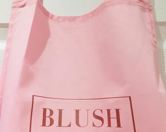 May include: A light pink apron with black adjustable straps. The apron features the word "BLUSH" in a brown rectangular frame, with "LIZ MCKENZI" and a scissors graphic below.