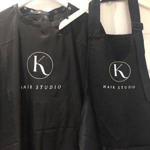 K&ouml;nnte beinhalten: Zwei schwarze Friseur-Sch&uuml;rzen mit einem wei&szlig;en Kreislogo mit dem Buchstaben K und dem Text "HAIR STUDIO".