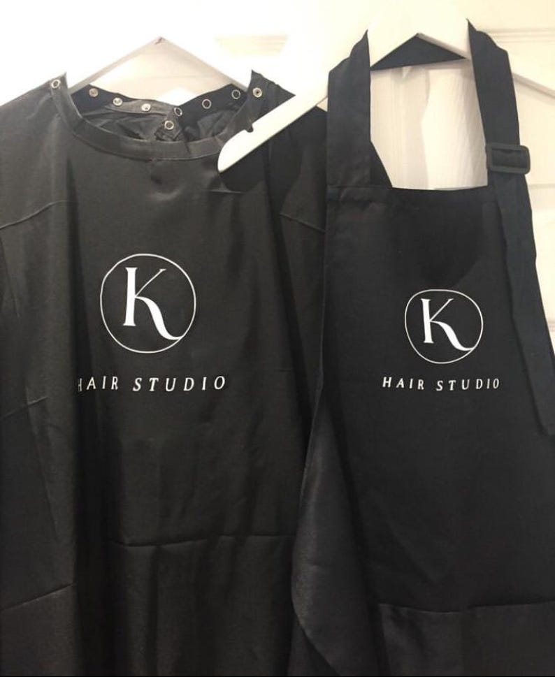K&ouml;nnte beinhalten: Zwei schwarze Friseur-Sch&uuml;rzen mit einem wei&szlig;en Kreislogo mit dem Buchstaben K und dem Text "HAIR STUDIO".