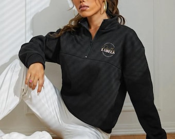 May include: Black quarter-zip sweatshirt with the text "LABELS" in a circular design. The sweatshirt has a high collar and a zipper closure. The model is wearing white pants and sneakers.