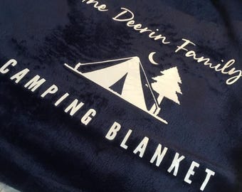 Könnte beinhalten: Marineblaue Campingdecke mit weißem Text und einer Camping-Szene-Grafik. Der Text lautet "The Deerin Family CAMPING BLANKET". Die Decke hat eine weiche Textur und ein weißes Sherpa-Futter.