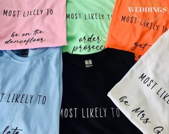 May include: Five different colored t-shirts with the text "Most Likely To" and a funny phrase on each shirt. The shirts are pink, green, orange, blue, and white.