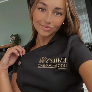 May include: Black dog grooming uniform with gold text reading "Emily's Dog Grooming". The uniform features a button closure and a collared neckline. The image shows a person wearing the uniform.