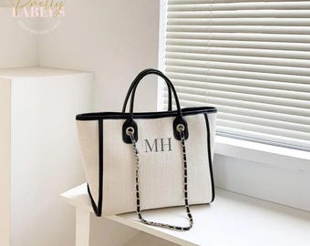 May include: A white canvas tote bag with black trim and a silver chain strap. The bag has the initials "MH" embroidered on the front.