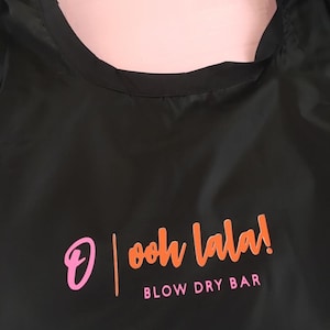 K&ouml;nnte beinhalten: Schwarze wiederverwendbare Einkaufstasche mit dem Text "ooh la la!" und "BLOW DRY BAR" in orangefarbener und rosafarbener Schrift.